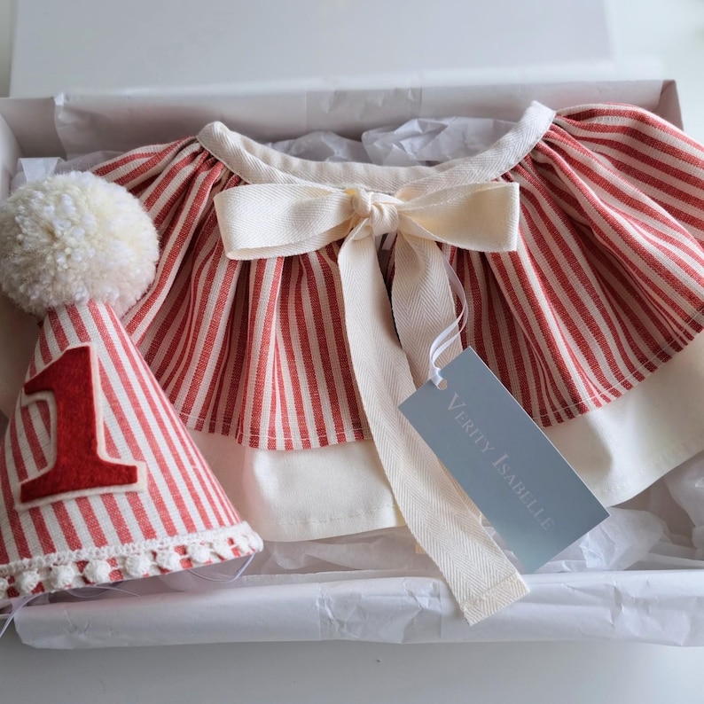 May include: A red and white striped outfit for a first birthday celebration, including a matching party hat with a large number 1. The outfit has a cream-colored bow and trim. The set is presented in a white box.