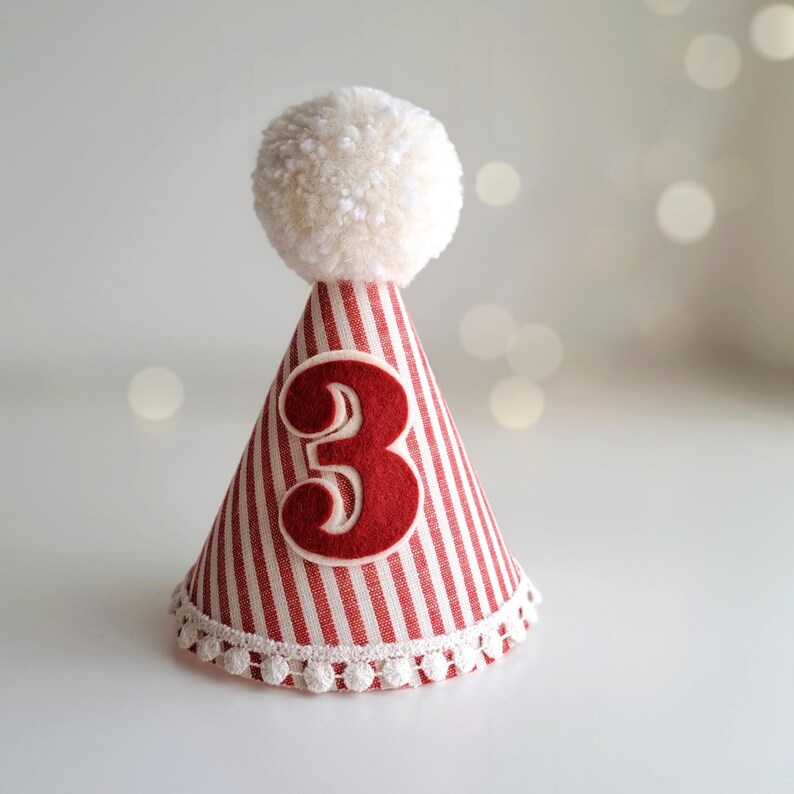 May include: A festive party hat with red and white stripes, a white pom-pom, and a large red number 3. The base is trimmed with white pom-poms. This birthday hat is perfect for a third birthday celebration.