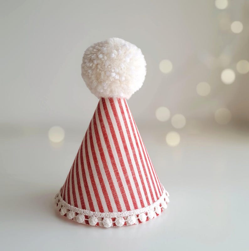 May include: A festive party hat with red and white stripes, topped with a fluffy white pom-pom. The base of the hat is trimmed with a delicate white pom-pom lace. This decorative hat is perfect for celebrations.