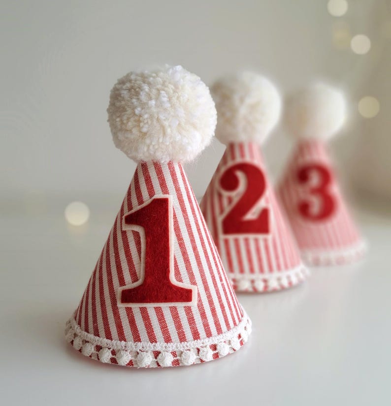 May include: Three red and white striped party hats with white pom-poms on top. The hats are numbered 1, 2, and 3 in red. The base of each hat is trimmed with white lace.
