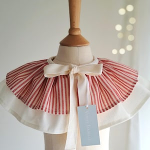 May include: A red and white striped collar with a cream-colored bow and trim. The collar is displayed on a mannequin head. A tag with the text "Verry Isabelle" hangs from the collar.
