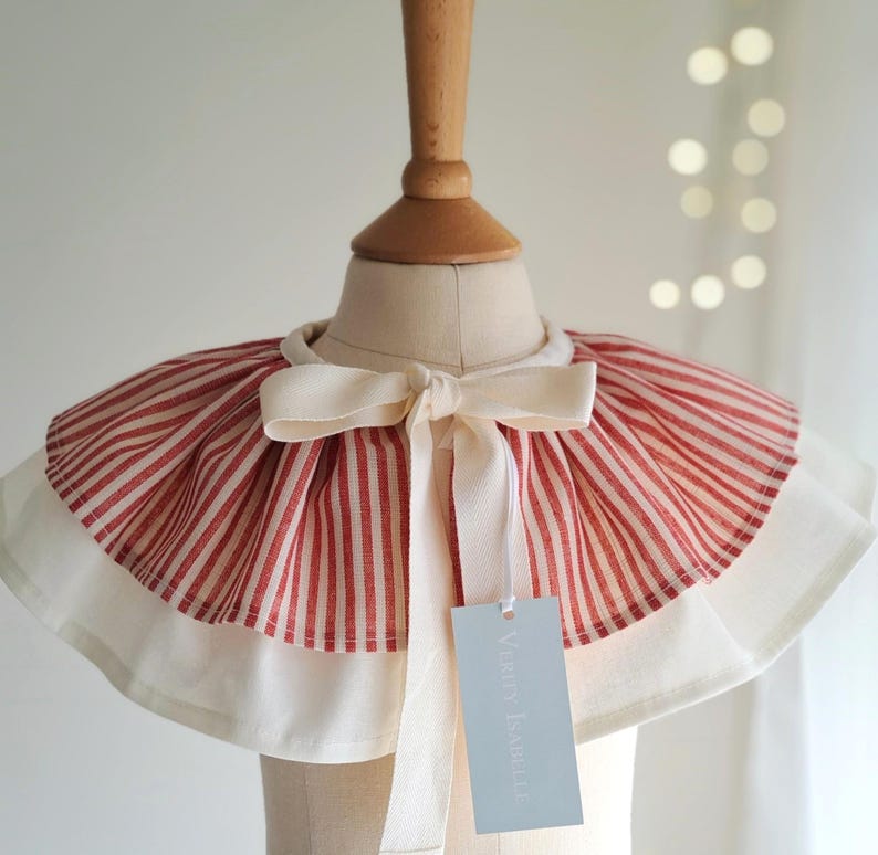 May include: A red and white striped collar with a cream-colored bow and trim. The collar is displayed on a mannequin head. A tag with the text "Verry Isabelle" hangs from the collar.