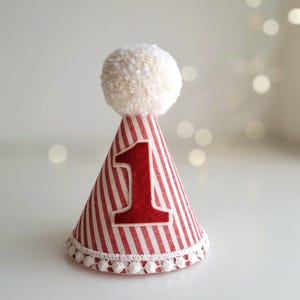 May include: A red and white striped party hat with a large red number 1 applique. The hat is topped with a fluffy white pom-pom and trimmed with white pom-poms at the base. Perfect for a first birthday celebration.