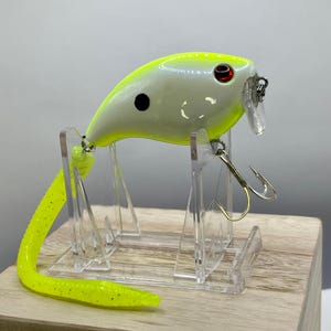 Pode incluir: Uma isca de pesca branca e verde chartreuse com cauda amarela e olhos vermelhos. A isca tem um ponto preto na lateral e é montada em um suporte de plástico transparente. Um anzol dourado é visível.