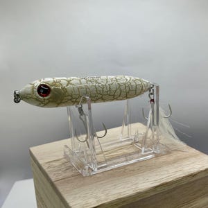 May include: A white and tan fishing lure with a cracked pattern, red eyes, and a silver hook. The lure has a white feather tail and is displayed on a clear acrylic stand. The lure is on a wooden block.