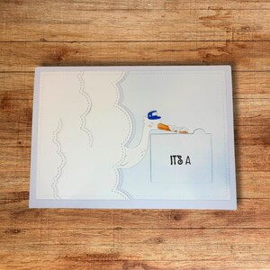 May include: A light blue greeting card with a white goose wearing a blue hat and carrying a carrot. The card has the text "IT'S A" on the front. The background is a wooden surface.