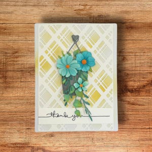 May include: A handmade greeting card featuring a hanging flower basket with turquoise flowers and green leaves. The background has a yellow and white plaid pattern. The words "thank you" are written in cursive.