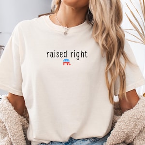 May include: Cream-colored t-shirt with the phrase "raised right" in black text. Below the text is a small graphic with red, blue, and white colors. The shirt is worn with blue jeans and a beige jacket.