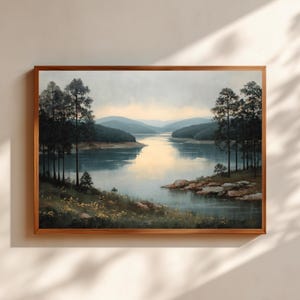 May include: A framed landscape painting depicts a serene lake scene. The artwork features a calm body of water reflecting the sky, surrounded by trees and rolling hills. The frame is a warm, natural wood tone.