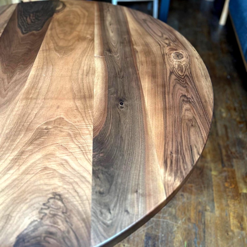 Puede incluir: Primer plano de una mesa redonda de madera con un acabado natural de vetas. La mesa tiene una combinaci&oacute;n de colores marr&oacute;n oscuro y claro, con nudos y patrones visibles en la madera. El borde de la mesa es redondeado.