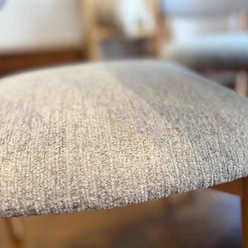 May include: Close-up of a chair seat with a textured, light beige fabric. The seat has a rounded edge and appears to be part of a wooden chair frame. The fabric has a woven appearance.