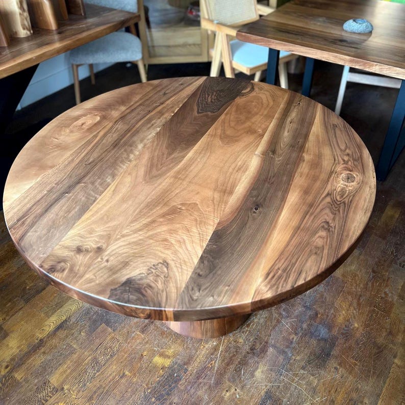 Puede incluir: Una mesa redonda de madera con un acabado de vetas naturales. La mesa tiene una base de pedestal y una superficie lisa y pulida. Los tonos de madera var&iacute;an de marr&oacute;n claro a oscuro, creando una est&eacute;tica c&aacute;lida. La mesa es adecuada para comer o como pieza decorativa, con unos 100 cm de di&aacute;metro.