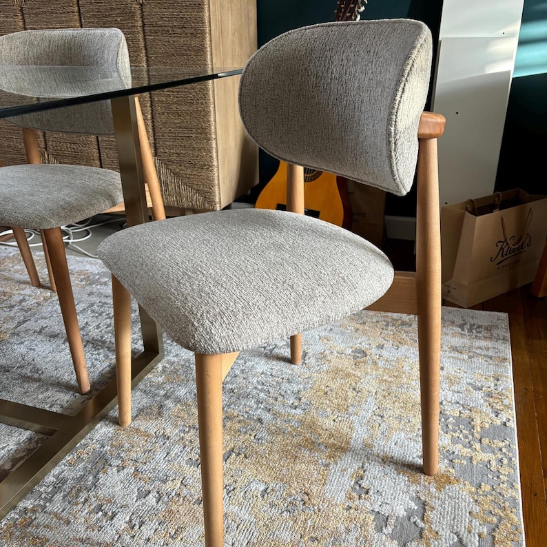 May include: Two light gray upholstered chairs with wooden legs and backs. The chairs have a mid-century modern design. The chairs are placed on a patterned rug. The chairs are near a glass table.