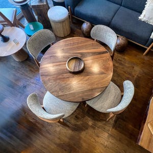 May include: A round wooden dining table with a dark finish and a central recessed area. Four chairs with gray fabric seats and curved backs surround the table. The chairs have wooden legs. The table is surrounded by other furniture.