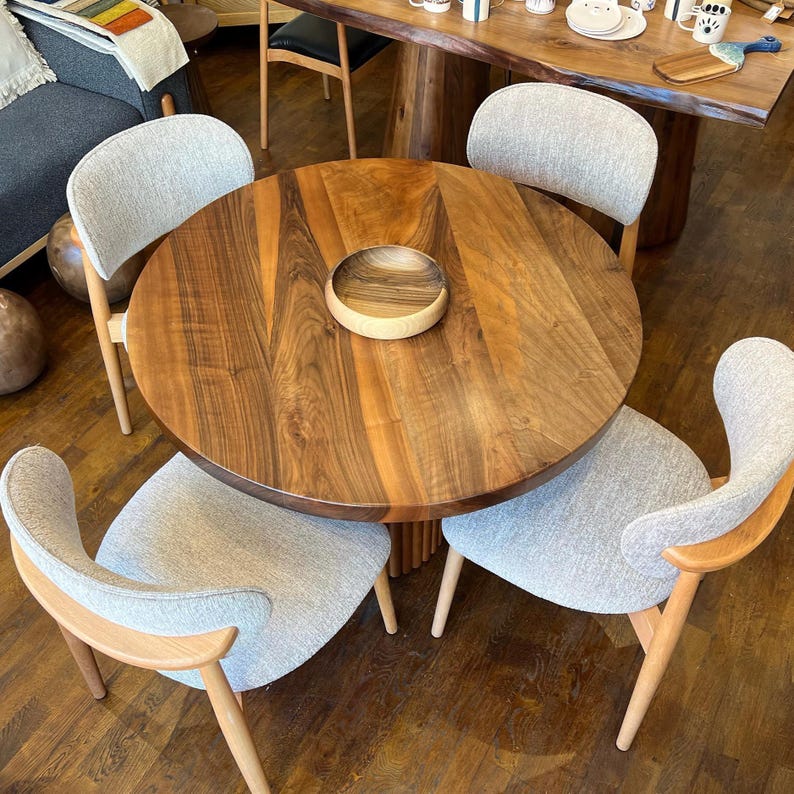 May include: A round wooden dining table with a natural wood grain finish, surrounded by four chairs with light gray upholstered seats and wooden frames. A small wooden bowl sits in the center of the table. The table is approximately 48 inches in diameter.