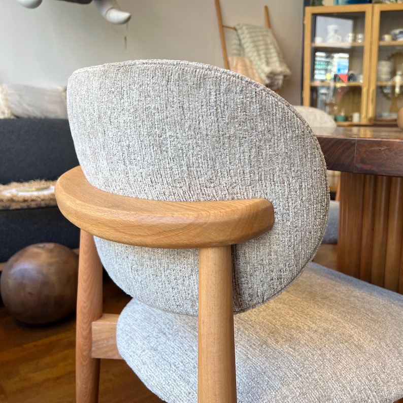 May include: A modern chair with a curved back and armrest. The chair features a light gray fabric seat and back, with a natural wood armrest and legs. The design is minimalist and the chair is suitable for a dining room or living room.
