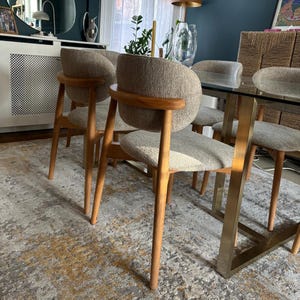 May include: A set of six dining chairs with light brown wooden legs and frames. The chairs have curved backs and seats upholstered in a neutral-toned fabric. The table has a glass top and gold-colored metal legs.