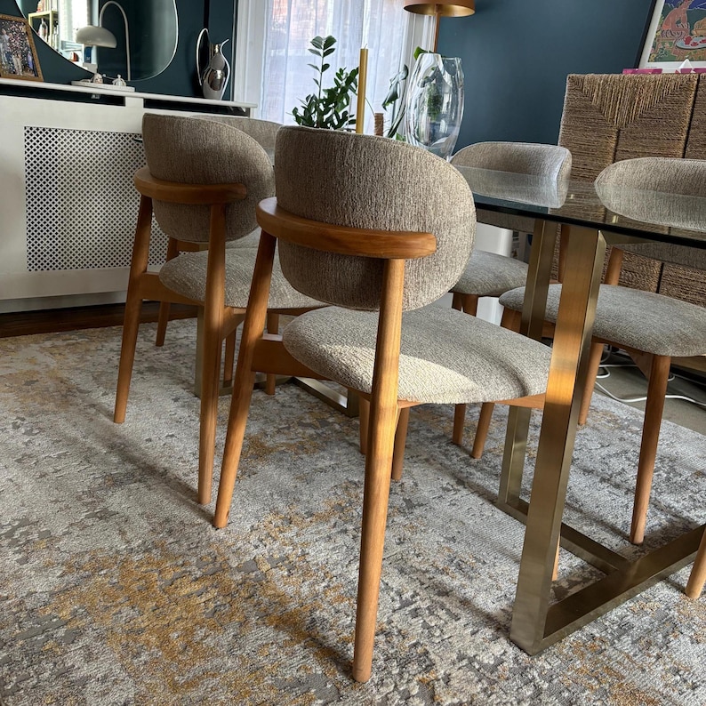 May include: A set of six dining chairs with light brown wooden legs and frames. The chairs have curved backs and seats upholstered in a neutral-toned fabric. The table has a glass top and gold-colored metal legs.