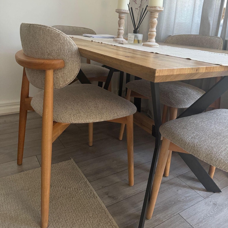 May include: A wooden dining table with a light-colored wood finish, paired with four upholstered chairs. The chairs have light-colored fabric seats and backs, with wooden legs and armrests. The table has black metal supports.