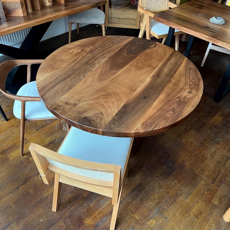 Puede incluir: Una mesa redonda de madera con un acabado de vetas naturales. La mesa est&aacute; rodeada de varias sillas con cojines de color claro y marcos de madera. El di&aacute;metro de la mesa es de aproximadamente 122 cm.