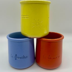May include: Three colorful ceramic jars stacked together. The top jar is yellow with embossed sun and wave designs. The bottom jars are blue and red, each with the script "la fermière". These jars are suitable for small plants or storage.