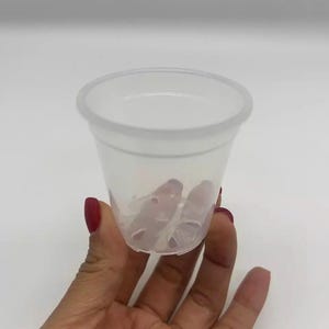 May include: A clear, translucent plastic cup with a rounded rim. The cup is held in a hand with red nail polish. The cup contains several clear, cylindrical objects.