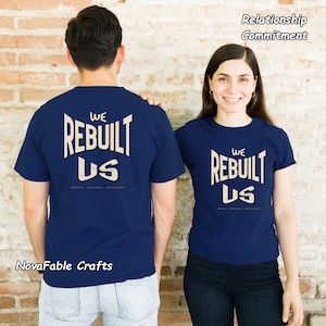Può includere: Due magliette blu navy con il testo bianco WE REBUILT US e le parole REPAIR RESTORE RECONNECT sottostanti. Il design è stampato sul retro di una maglietta e sul davanti dell'altra, con il testo Relationship Commitment ben visibile.