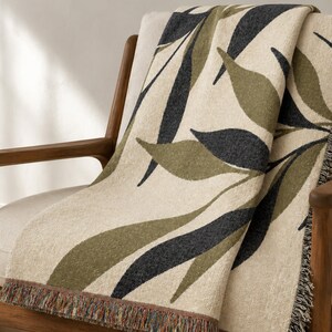 Olive woven cotton throw blanket draped over a wood-framed accent chair, featuring charcoal green and muted olive branch leaves on a warm cream field with visible fringe.