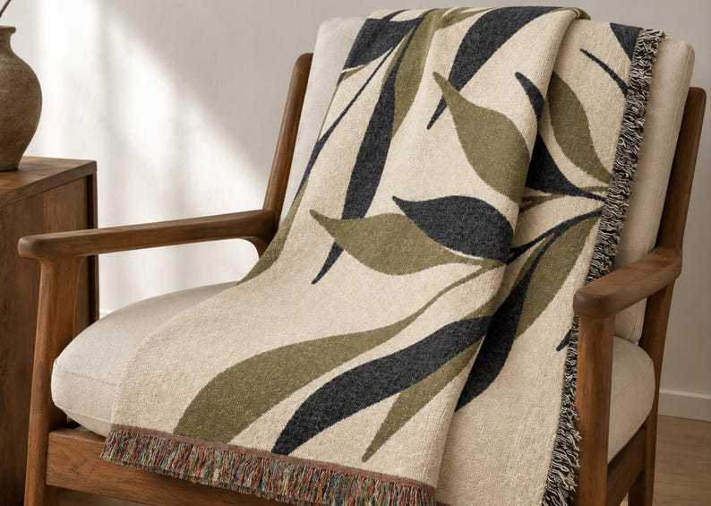 Olive woven cotton throw blanket draped over a wood-framed accent chair, featuring charcoal green and muted olive branch leaves on a warm cream field with visible fringe.