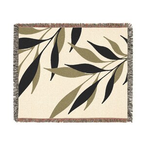Cream-coloured woven throw blanket featuring a repeating pattern of black and olive green leaves. The blanket has a fringed edge in shades of brown and tan. The leaves are stylized and arranged across the surface.