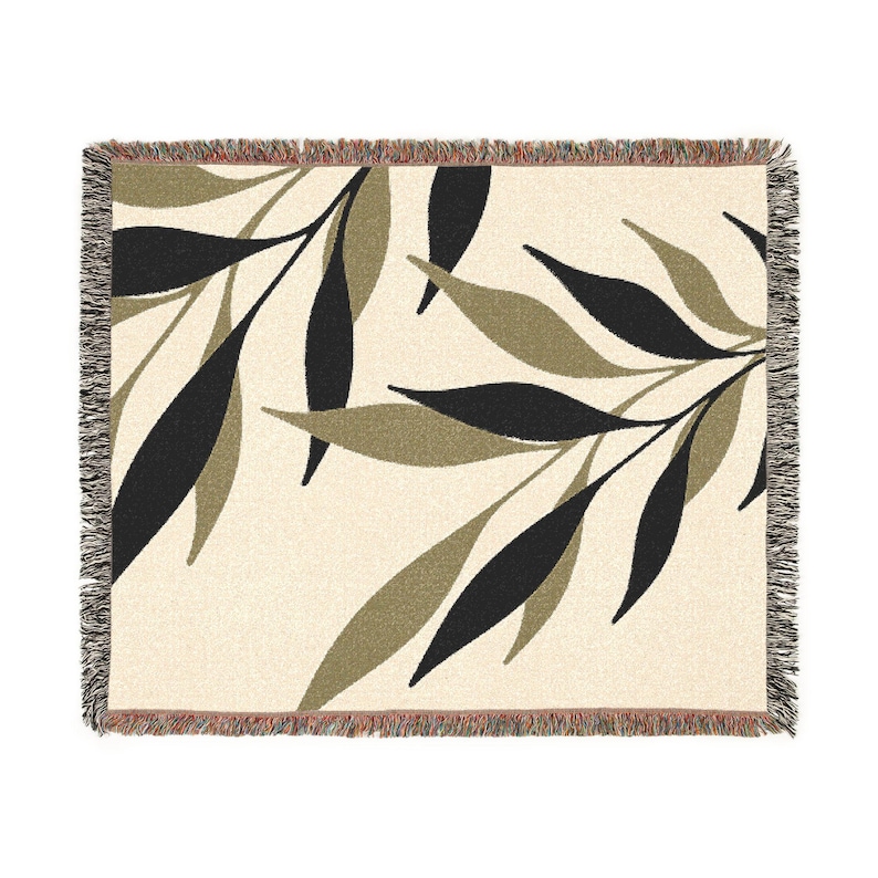 Cream-coloured woven throw blanket featuring a repeating pattern of black and olive green leaves. The blanket has a fringed edge in shades of brown and tan. The leaves are stylized and arranged across the surface.