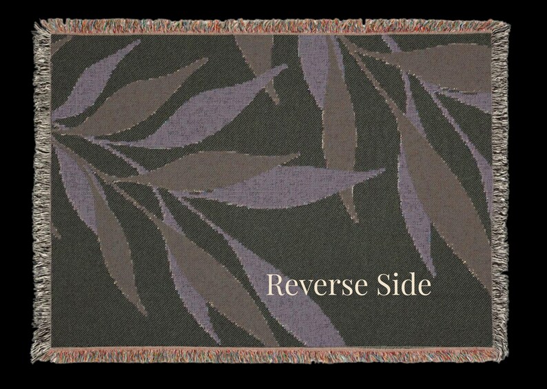 Reverse side of the Olive Branch woven cotton throw blanket laid flat, showing muted botanical leaves on a dark woven field with multi color fringe on all sides.