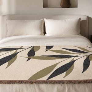 Olive woven cotton throw blanket laid across the foot of a neutral bed, featuring charcoal green and muted olive branch leaves on a warm cream field with fringed edges.