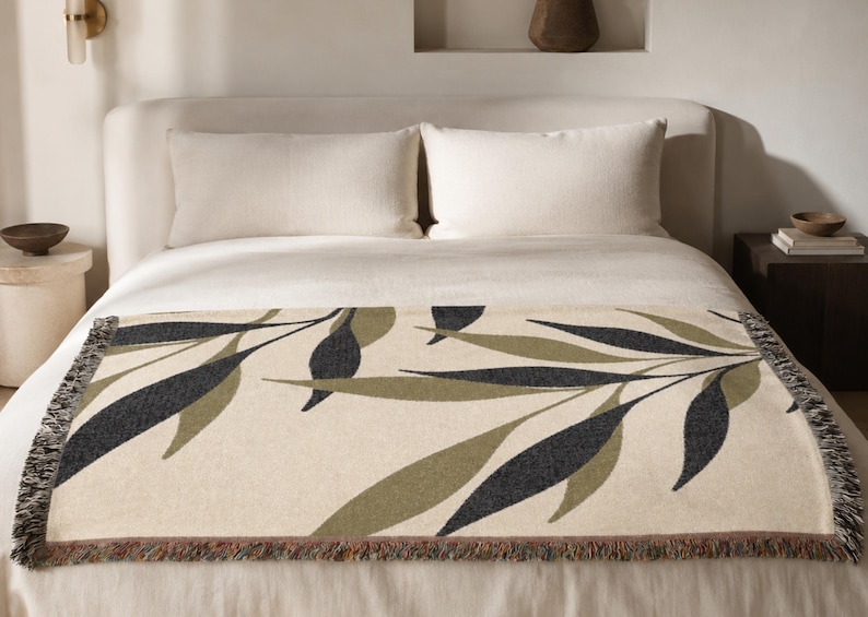 Olive woven cotton throw blanket laid across the foot of a neutral bed, featuring charcoal green and muted olive branch leaves on a warm cream field with fringed edges.