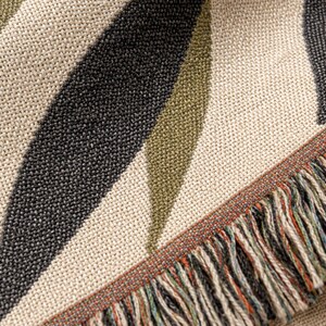 Close-up of the Olive Branch woven cotton throw blanket, showing muted olive and black leaf details, cream woven texture, and multi color fringe edge.