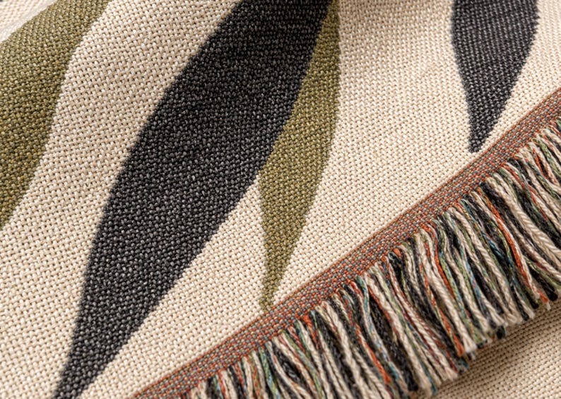Close-up of the Olive Branch woven cotton throw blanket, showing muted olive and black leaf details, cream woven texture, and multi color fringe edge.