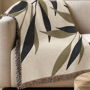 Olive woven cotton throw blanket draped over a neutral sofa, featuring charcoal green and muted olive branch leaves on a warm cream field with visible fringe.
