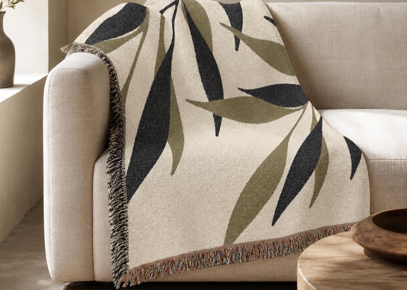 Olive woven cotton throw blanket draped over a neutral sofa, featuring charcoal green and muted olive branch leaves on a warm cream field with visible fringe.