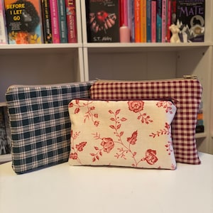 May include: Three fabric pouches with different patterns. One is dark green plaid, another is cream with red floral designs, and the third is red and white gingham. All have gold zippers. Bookshelves are in the background.