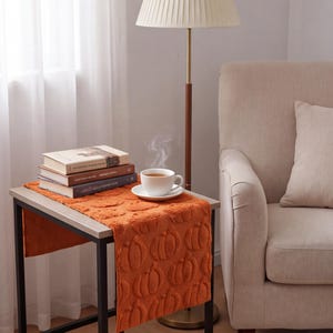 Peut inclure: Un chemin de table orange rectangulaire avec un motif de citrouilles en relief texturé, posé sur une table d'appoint. Une pile de trois livres et une tasse blanche avec sa soucoupe contenant une boisson chaude fumante sont posées dessus.