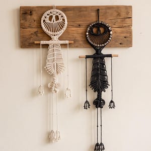 May include: Two macrame wall hangings featuring a human-like figure design. One is cream-colored and the other is black. Each piece consists of a circular head, a horizontal wooden dowel for shoulders, and a long, intricate knotted body structure.