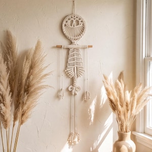 May include: A cream-colored macrame wall hanging featuring a circular top section with intricate knotting, suspended from a horizontal wooden dowel. The design includes a central vertical element with layered fringe and two long hanging cords.