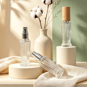May include: Three clear glass bottles with vertical ribbing and silver spray nozzles. One bottle stands upright on a round pedestal, another lies horizontally, and the third features a light wood cap, positioned near a vase with cotton stems.