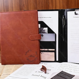 Könnte beinhalten: Ein braunes Leder-Portfolio mit Schnallenverschluss, personalisiert mit dem Namen "Alticus" in einem Lorbeerkranz. Das Portfolio ist geöffnet und zeigt ein schwarzes Innere mit Taschen, einem Stift und Dokumenten.