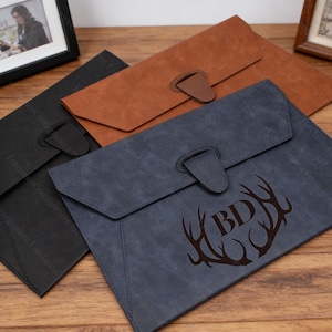 May include: Three leather document holders in black, brown, and blue. Each has a flap closure with a tab and loop. The blue holder features a deer antler design with the initials "BD".