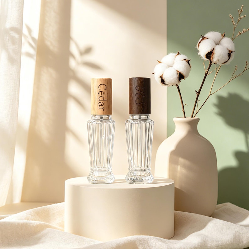 May include: Two clear glass spray bottles with vertical ribbed textures and cylindrical wooden caps. The left cap is light wood with the word Cedar engraved, and the right cap is dark wood with an engraved script monogram.