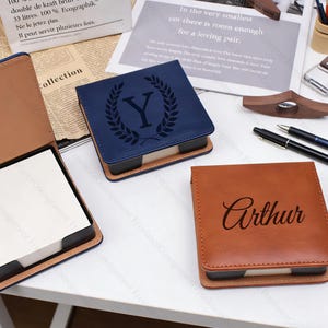 Puede incluir: Blocs de notas de cuero personalizados en azul marino y marrón. El bloc azul marino presenta una corona de laurel y la letra "Y", el marrón el nombre "Arthur". Cada bloc es cuadrado y tiene un inserto de papel claro.