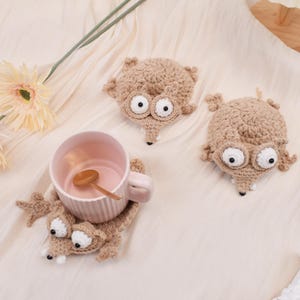 Peut inclure: Trois dessous de verre faits à la main en crochet en forme de créature de dessin animé avec de grands yeux blancs et un long museau. Un dessous de verre contient une tasse rose avec une cuillère en bois. Les dessous de verre sont beiges.