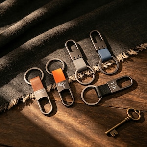 Puede incluir: Una colección de llaveros con correas de cuero en varios colores: marrón, naranja, gris, azul y negro. Cada llavero tiene un cierre metálico y un anillo metálico. Un llavero tiene las iniciales "TE" grabadas.