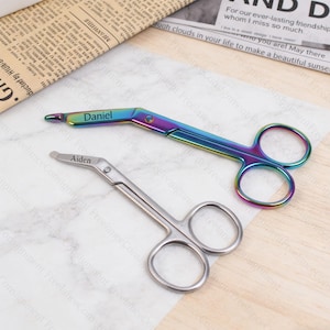May include: Two pairs of scissors. One pair is silver, and the other is iridescent with the name "Daniel" engraved on it. The silver pair has the name "Aiden" engraved on it. Both pairs are on a light-colored surface.